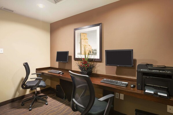 A hotel room with a desk, two monitors, a printer, a keyboard, a chair, and a framed picture on the wall.