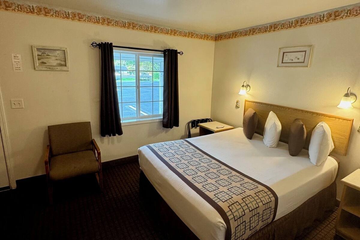 A hotel room with a large bed, a chair, a desk, and a window with curtains.