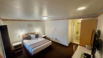 A hotel room with a bed, a desk with a computer, a chair, a TV, and a bathroom door.