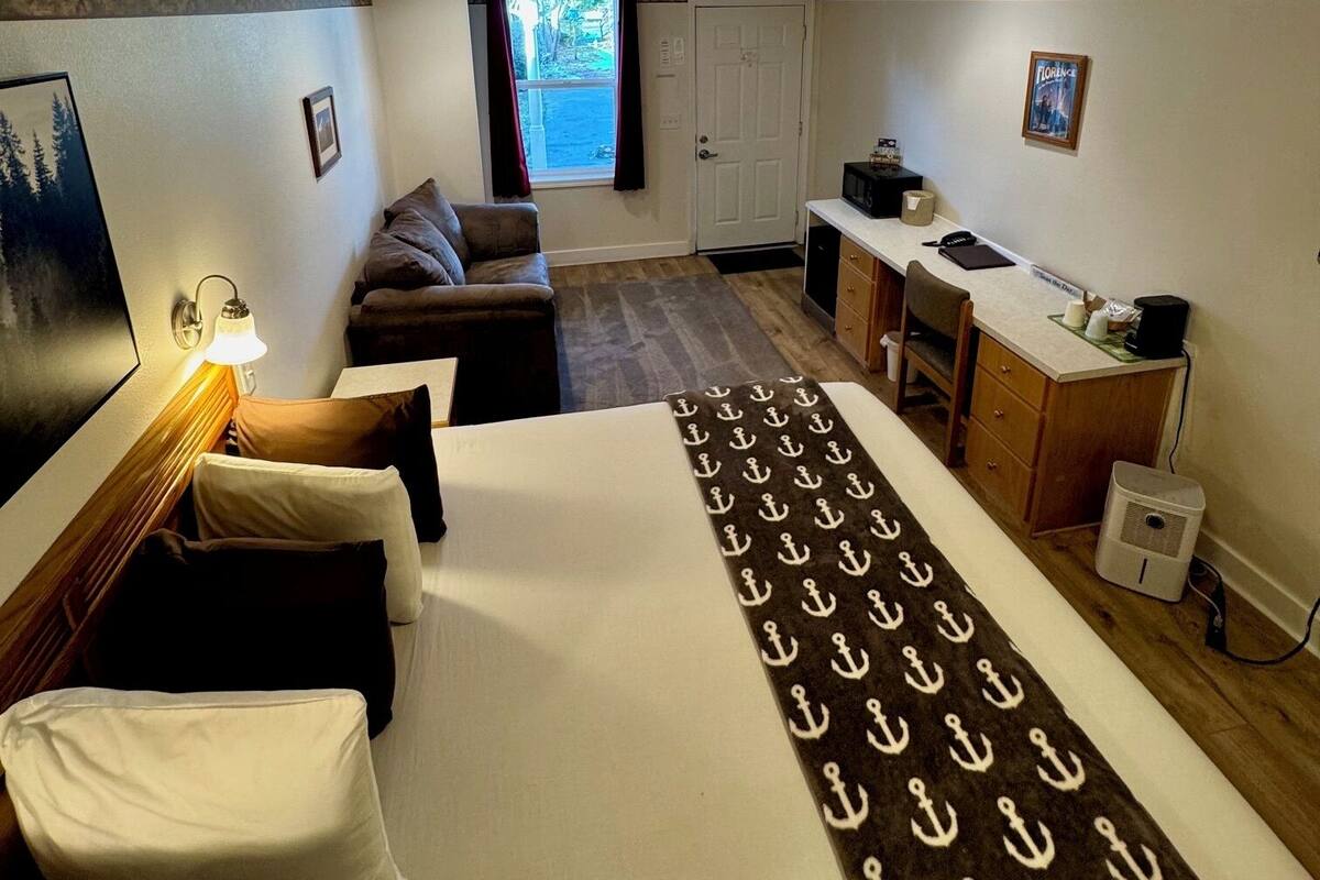 A compact hotel room with a bed, a small desk, a kitchenette, and a window with curtains.