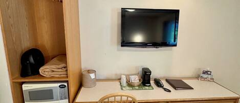 Ein kompaktes Hotelzimmer mit Schreibtisch aus Holz, einem an der Wand befestigten Fernseher, einer Mikrowelle, einem kleinen Kühlschrank und einem Sessel.
