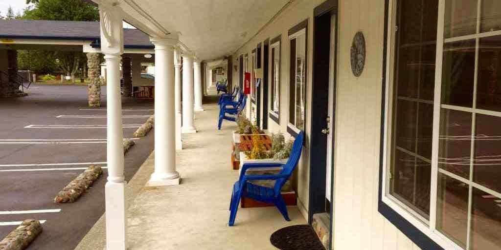 Un espacio al aire libre cubierto con una fila de sillas azules y una mesa, un estacionamiento y un edificio con grandes ventanales.