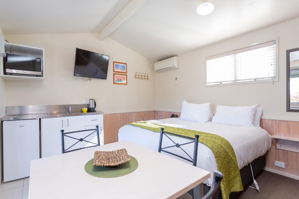 Quarto de hotel compacto com cozinha compacta, uma cama com manta verde, TV de tela plana e janela com persianas.