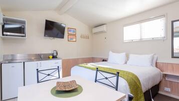 Quarto de hotel compacto com cozinha compacta, uma cama com manta verde, TV de tela plana e janela com persianas.