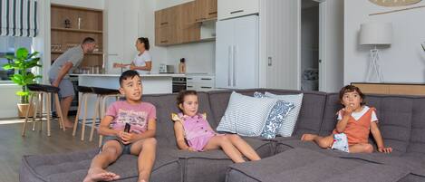 Una sala de estar moderna con un sofá seccional gris, una cocina al fondo y un comedor con sillas de madera.