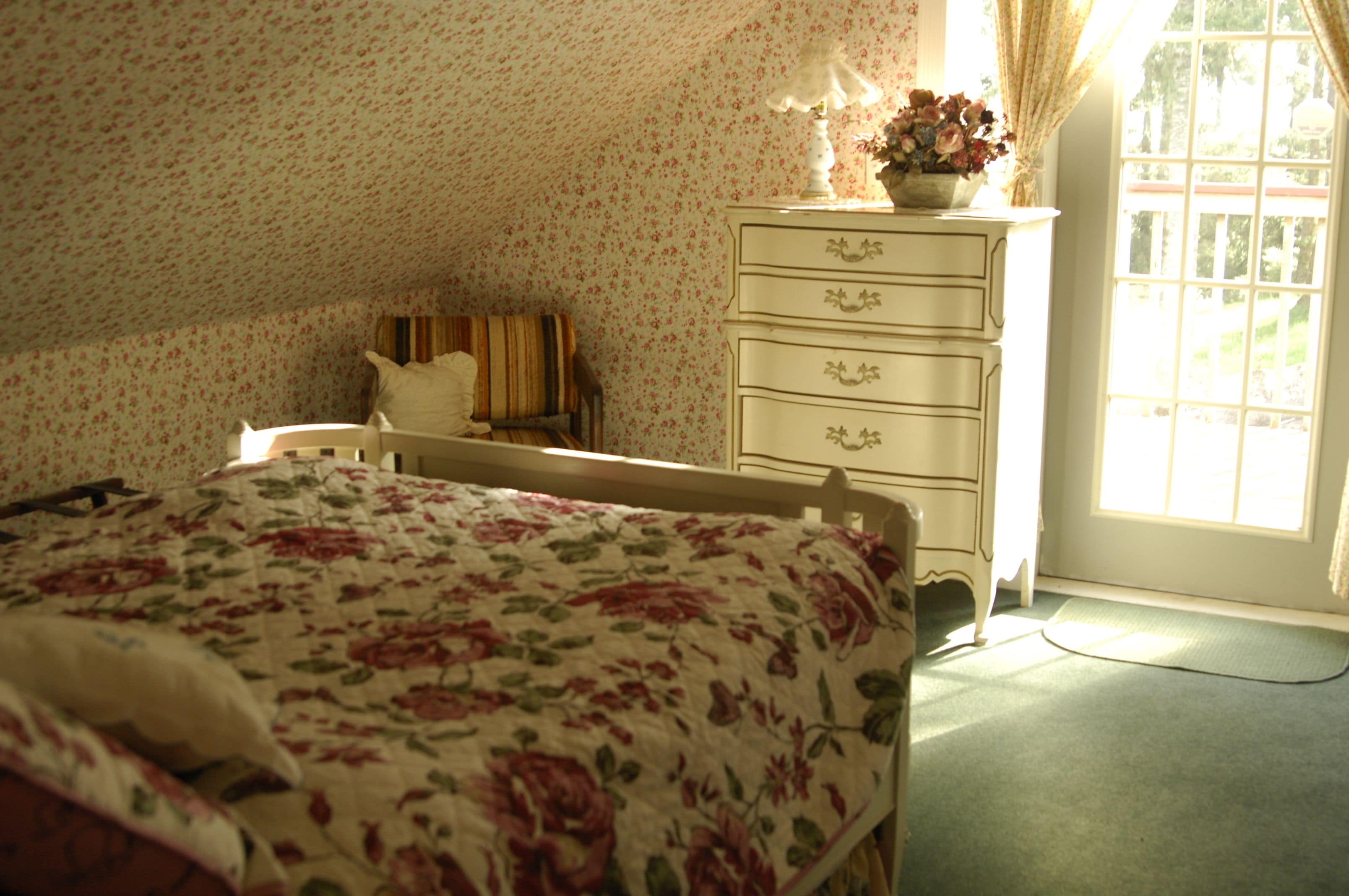 Una camera da letto con carta da parati a fiori, un letto con copripiumino floreale, una cassettiera e una finestra con tende.