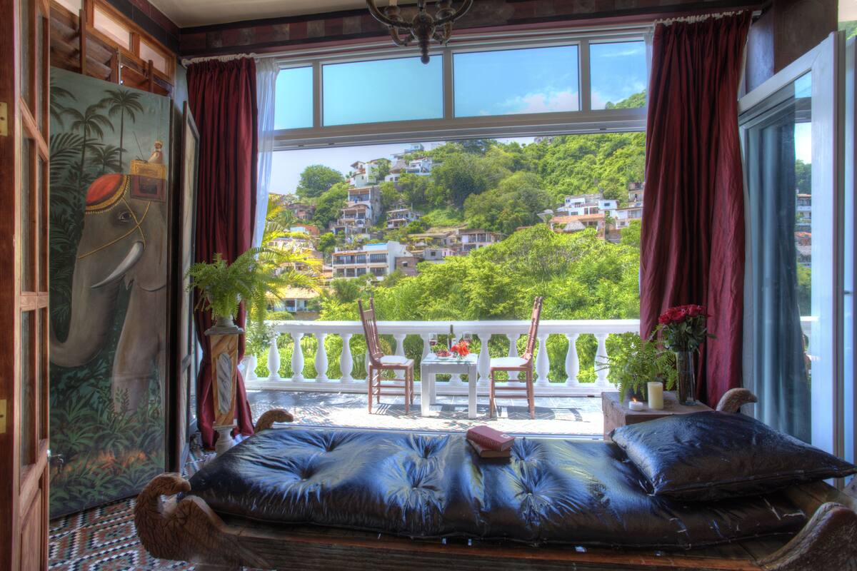 A room with a large window, a balcony, and a view of a hillside.