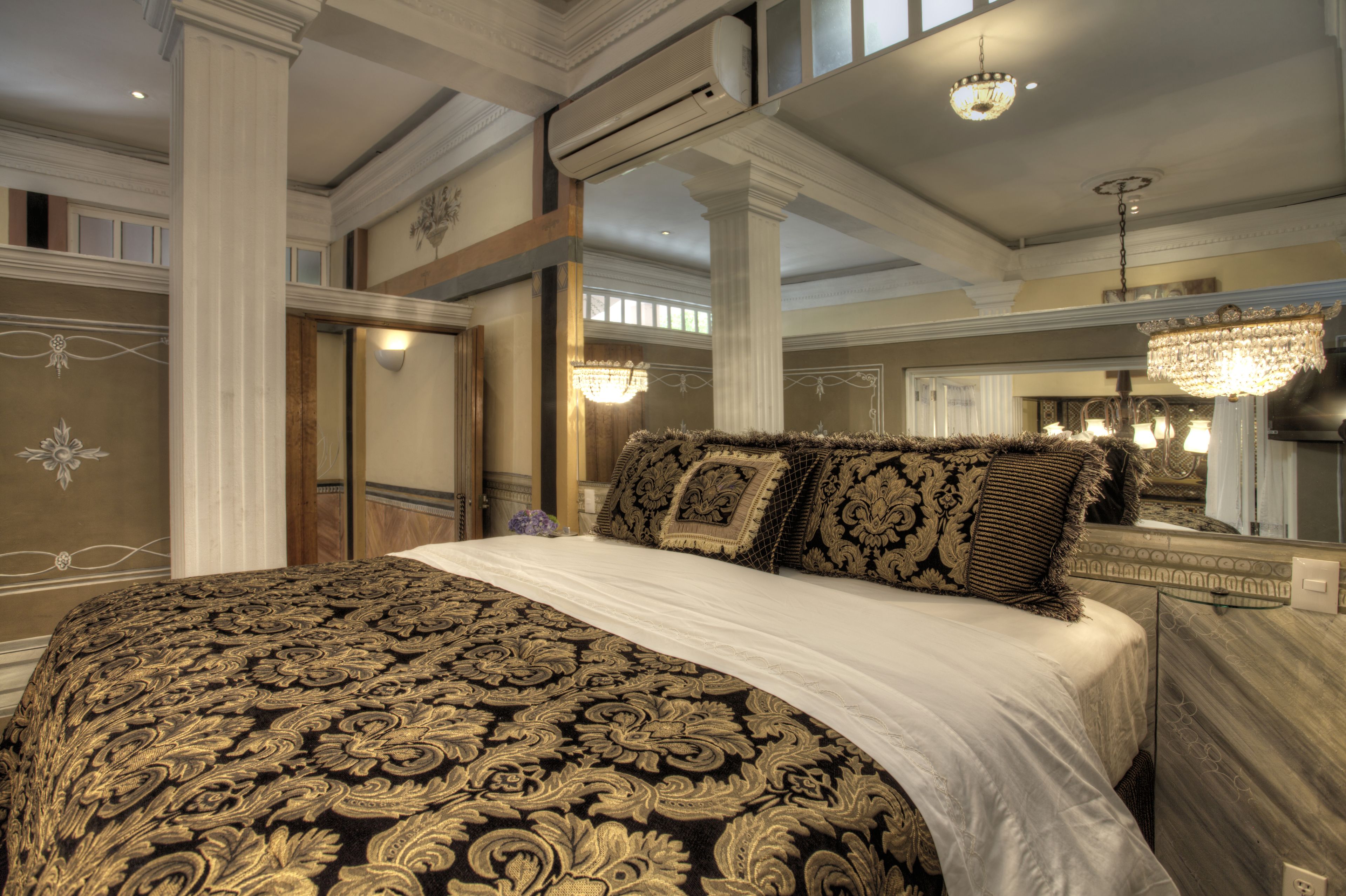 Una cama grande con ropa de cama decorada, una araña y un espejo grande en una habitación bien iluminada con techos altos y detalles arquitectónicos clásicos.