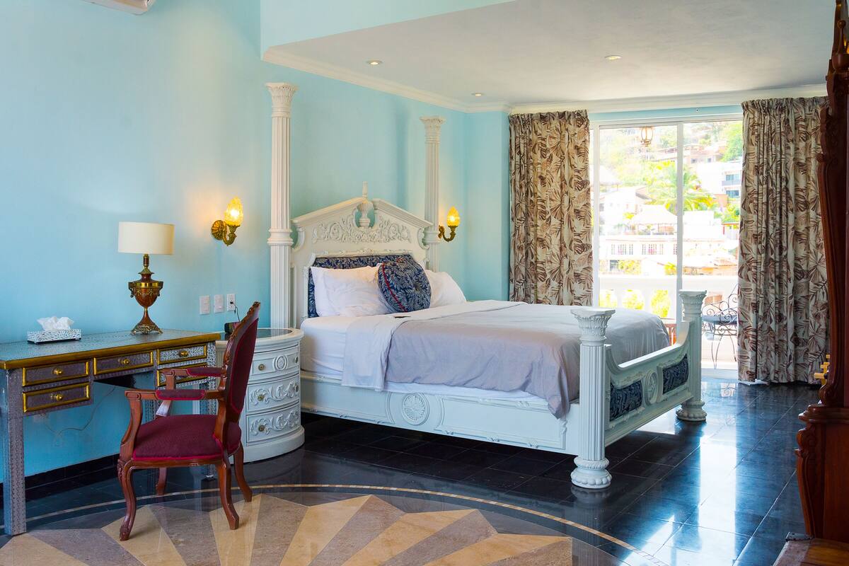 A bedroom with a large bed, a desk with a lamp, a chair, and a view of the outside.