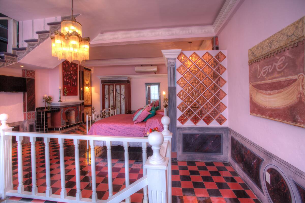 A bedroom with a checkered floor, a bed with pink bedding, a chandelier, and a fireplace.