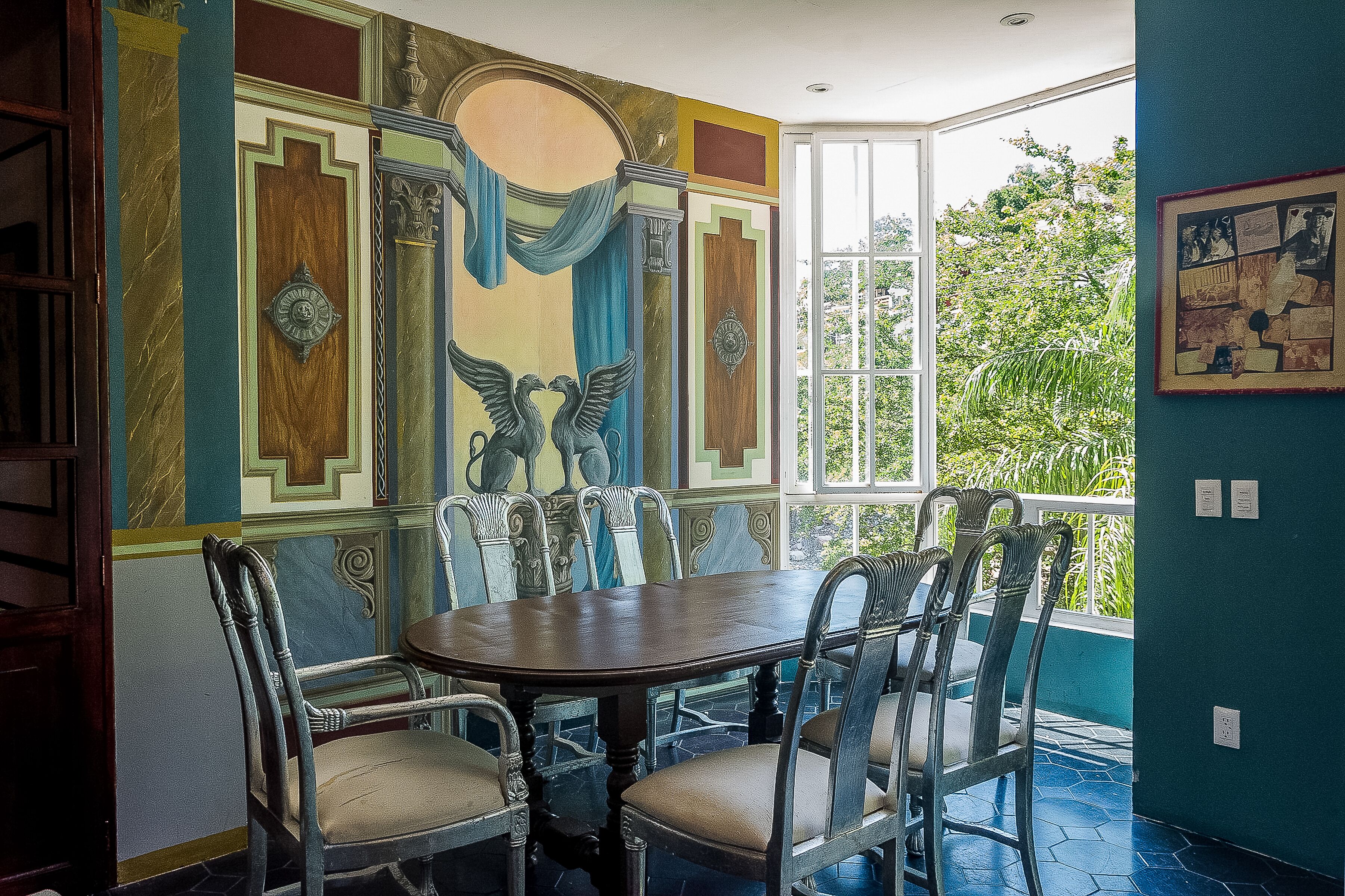 Een eetgedeelte met een ronde tafel en zes stoelen, een muurschildering met geometrische patronen en een centrale figuur, en uitzicht op groen door een groot raam.