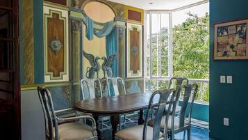 Een eetgedeelte met een ronde tafel en zes stoelen, een muurschildering met geometrische patronen en een centrale figuur, en uitzicht op groen door een groot raam.