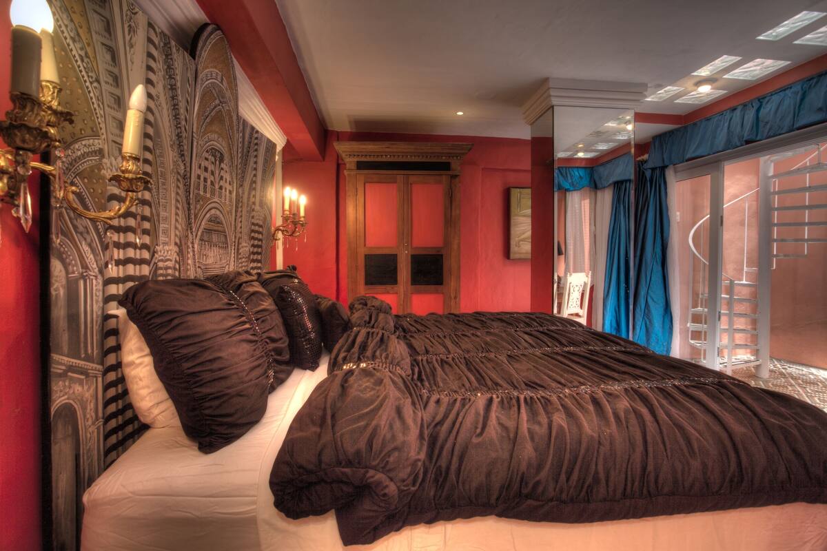 A large bed with a dark brown bedspread, a chandelier, and a red wall with a detailed mural.
