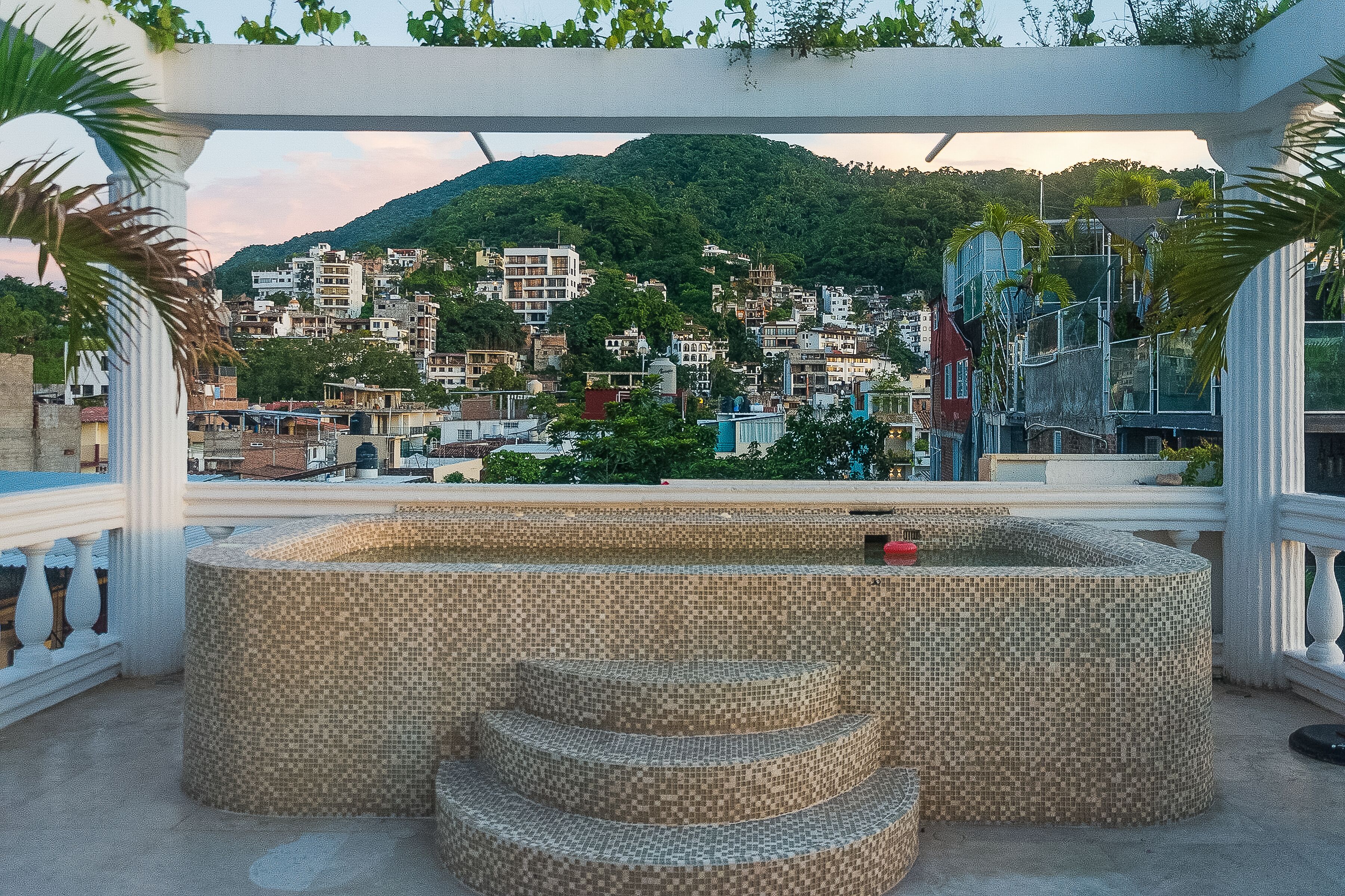 Un área de estar exterior con baldosas y escalones, rodeada por una pérgola blanca.