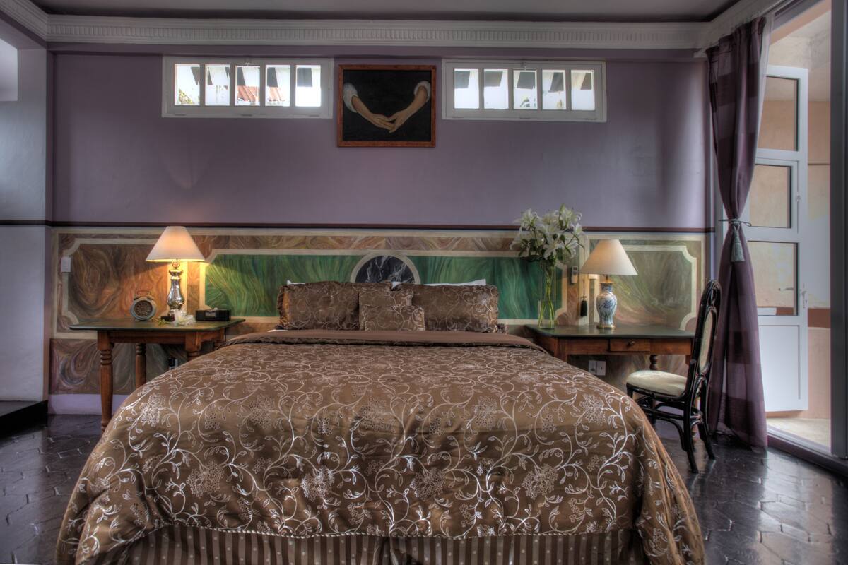 A bedroom with a large bed, two bedside tables with lamps, a painting above the bed, and a window with curtains.