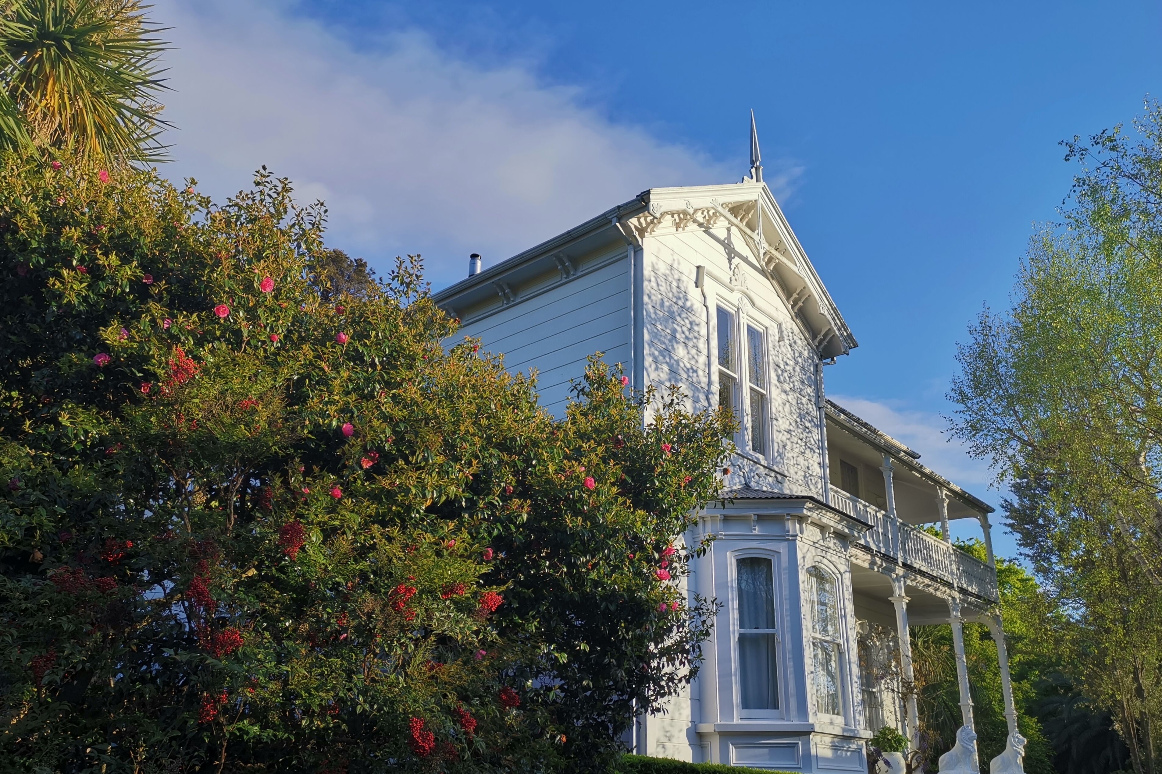 A white building with a balcony and columns, surrounded by lush greenery and red flowers.