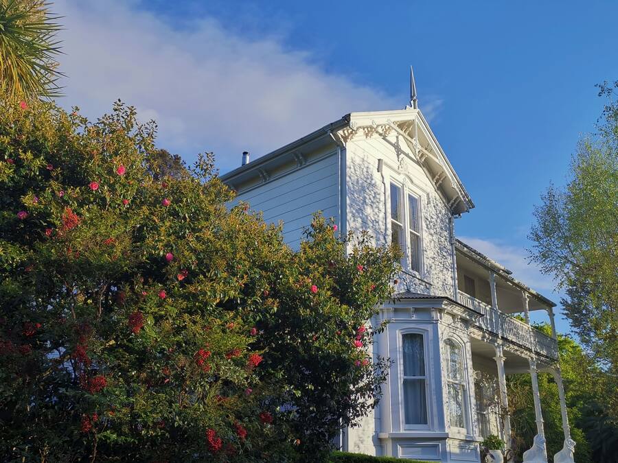 A white building with a balcony and columns, surrounded by lush greenery and red flowers.