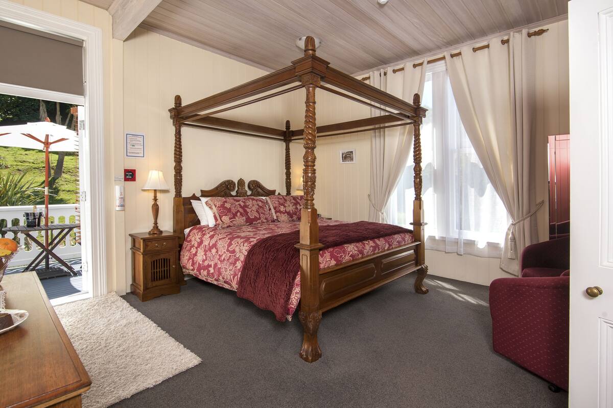 A four-poster bed with a floral bedspread, a wooden nightstand with a lamp, and a red armchair in a bedroom with a view of the outdoors.