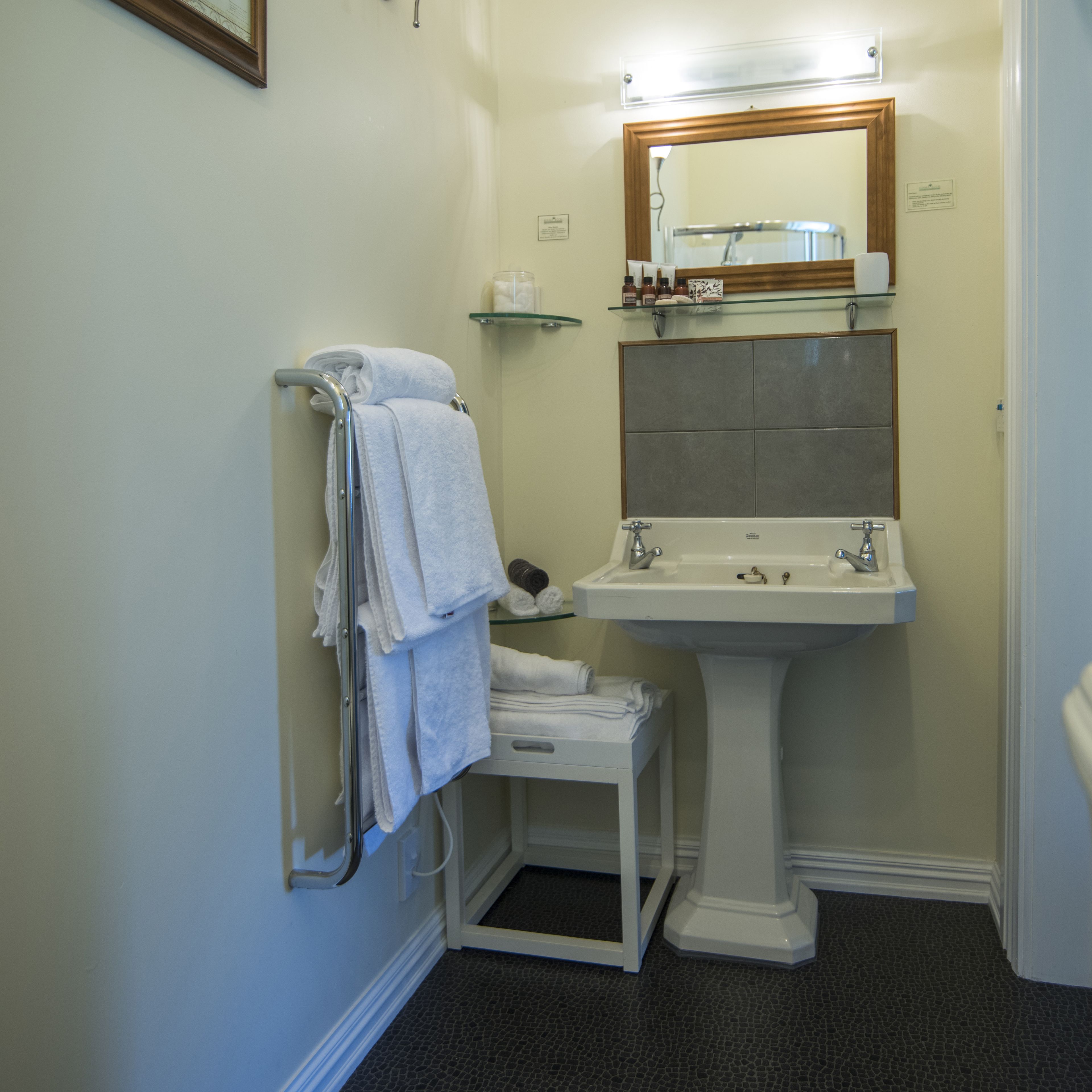 Un baño con un lavamanos pedestal blanco, un estante de vidrio sobre el lavamanos, y un perchero con toallas dobladas.