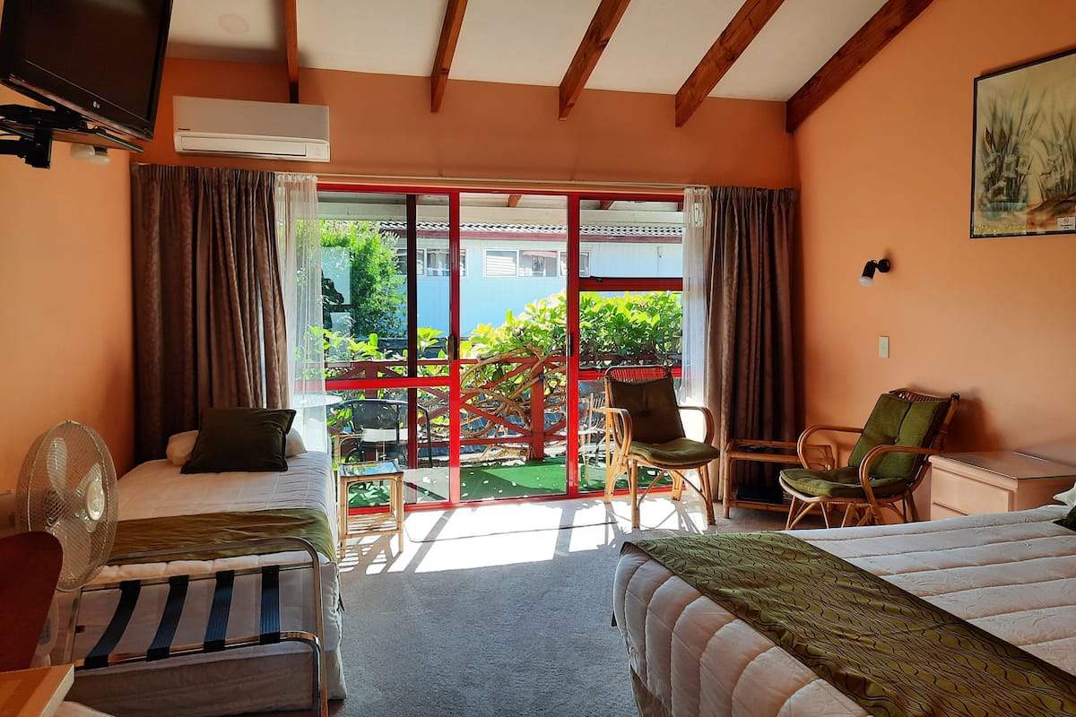 A hotel room with two beds, a desk, a chair, a fan, and a view of the outside.
