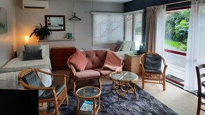 Un salon confortable avec un lit, un canapé, une table basse et un fauteuil. Il y a une fenêtre avec des stores, un luminaire suspendu et une plante en pot.