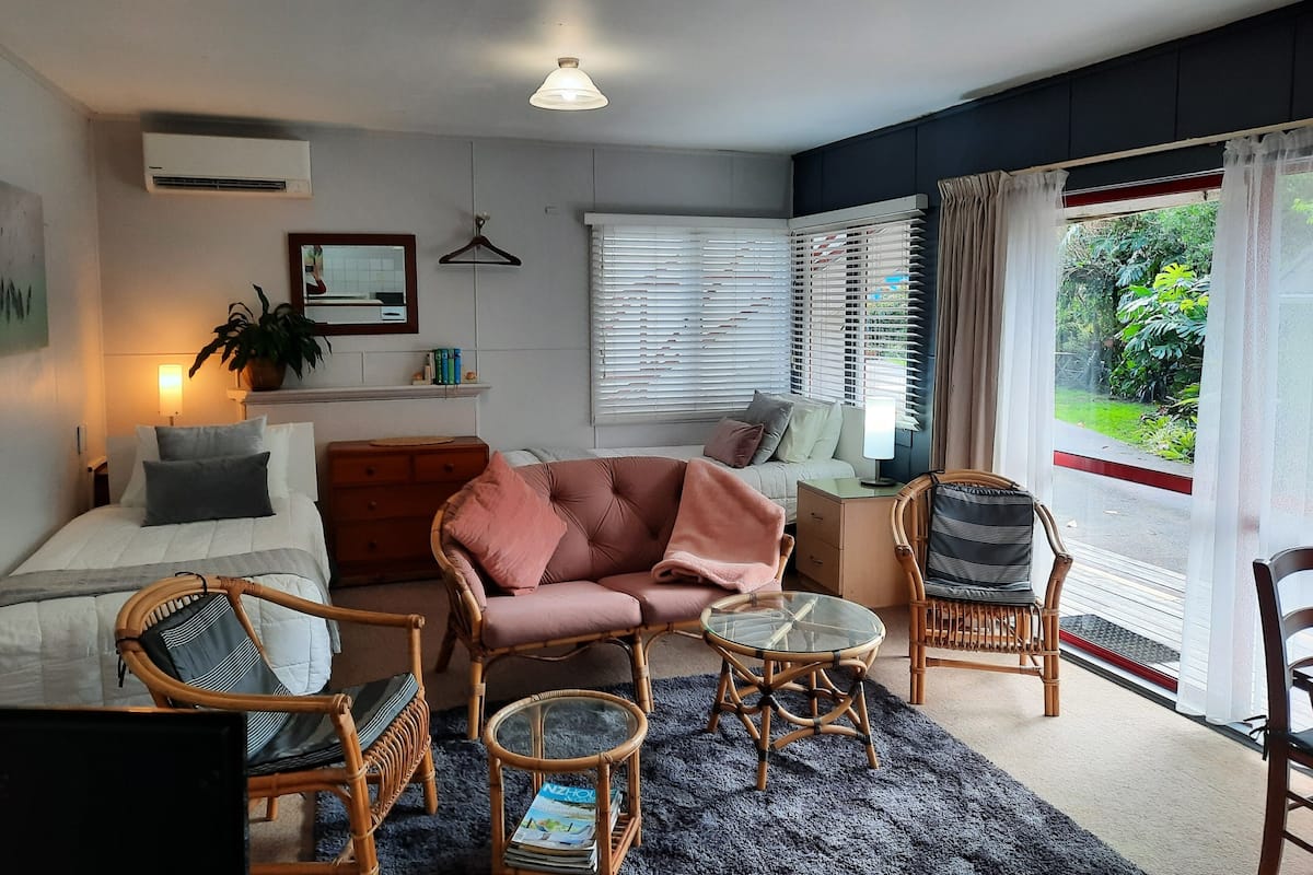 A cozy living room with a bed, a sofa, a coffee table, and a chair. There is a window with blinds, a hanging light fixture, and a potted plant.