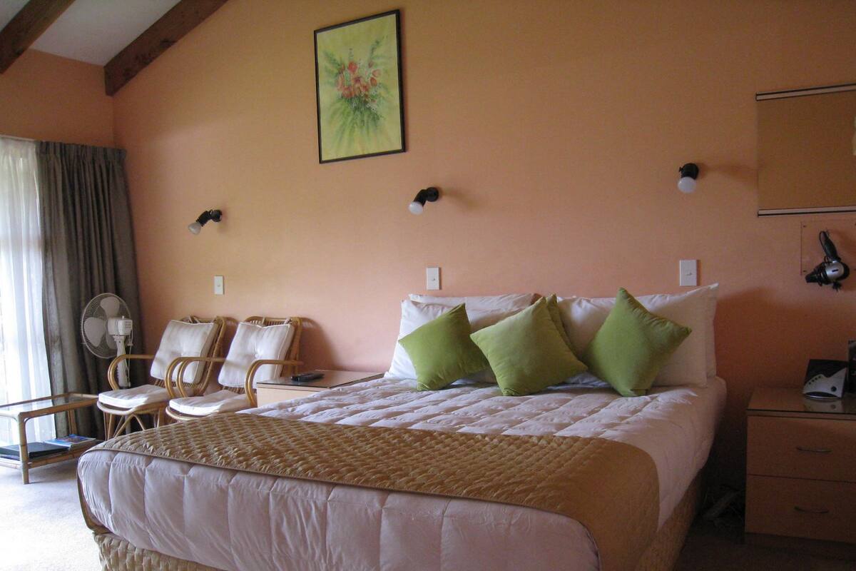 A hotel room with a large bed, two chairs, a desk, and a fan.