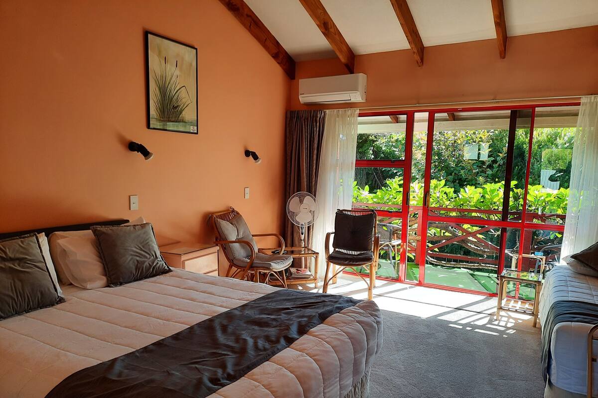 A bedroom with a large bed, a desk, two chairs, a fan, and a view of greenery through a glass door.