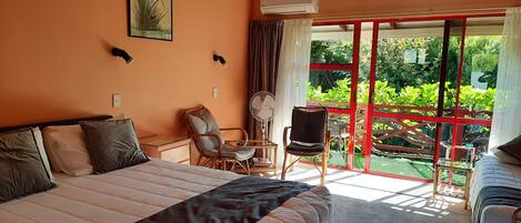 A bedroom with a large bed, a desk, two chairs, a fan, and a view of greenery through a glass door.
