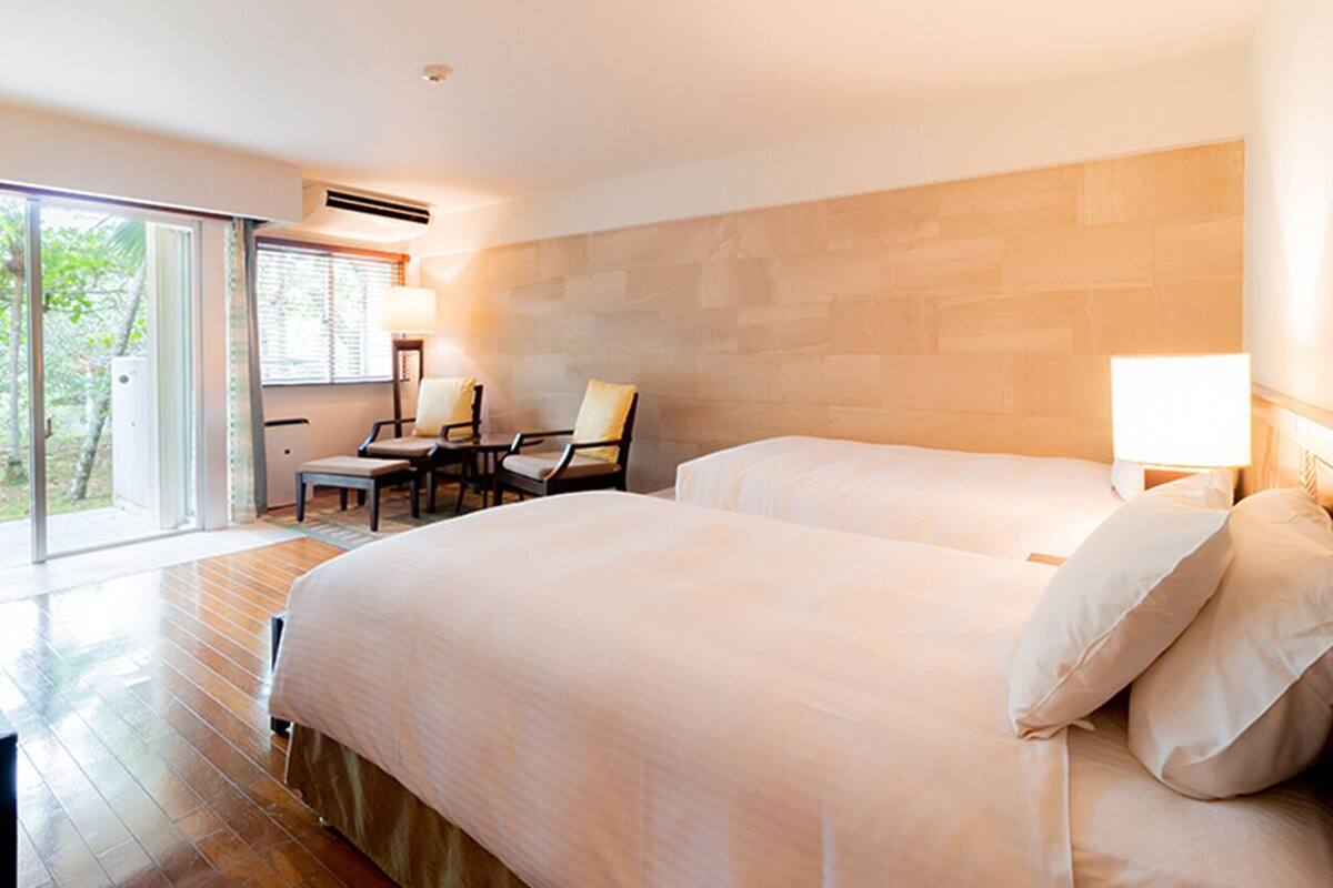 A modern hotel room with a large bed, two chairs, a desk, and a sliding glass door leading to a balcony.