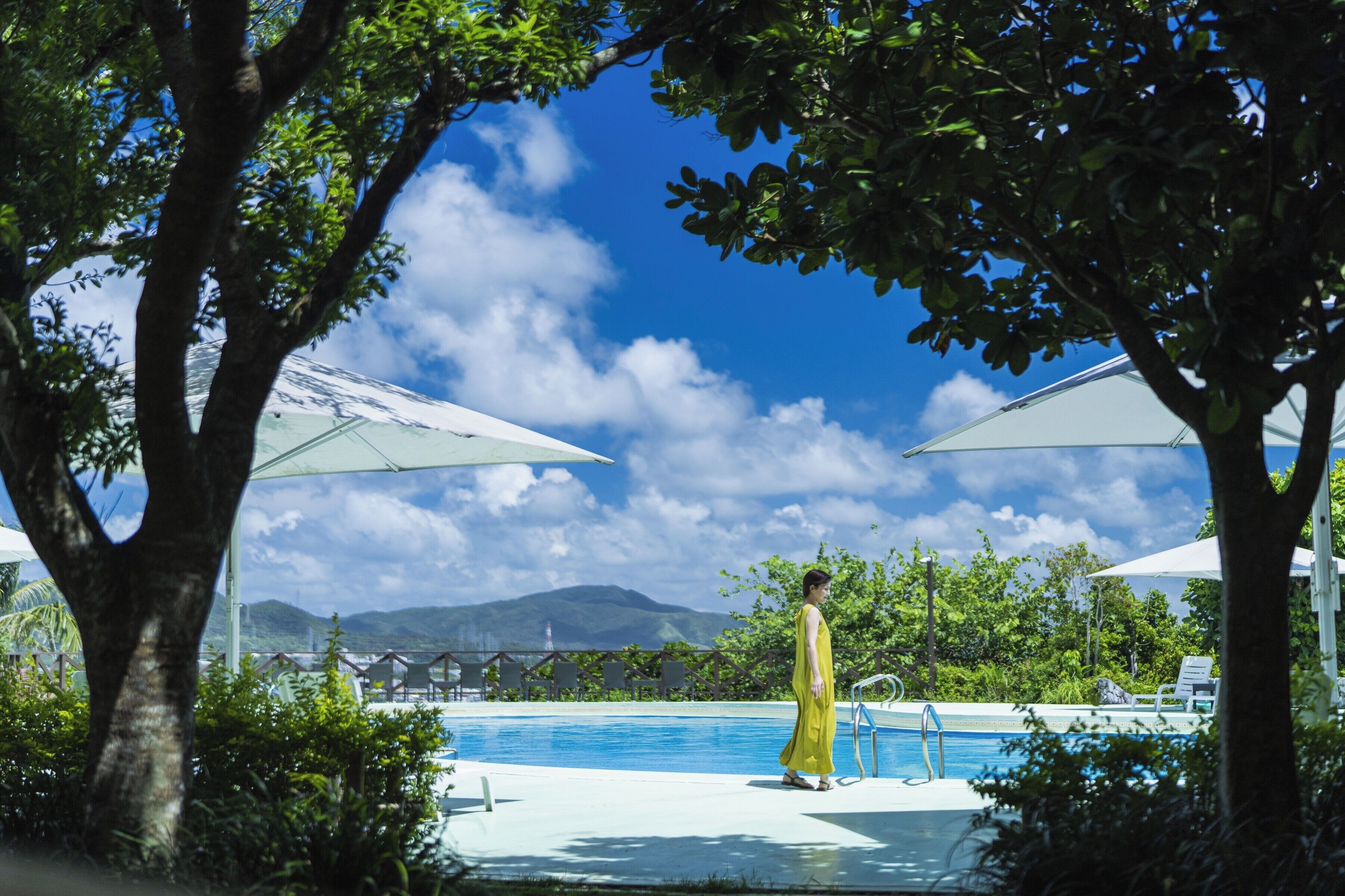 A pool area with a person in a yellow dress, trees, and a clear sky.