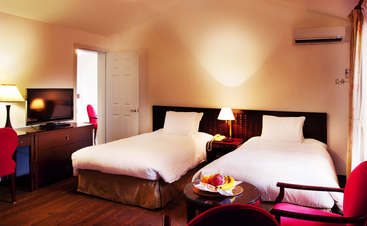 Ein Hotelzimmer mit zwei Betten, einem Schreibtisch mit Fernseher, einem Sessel, einem Nachttisch mit Lampe und einer Obstschale.