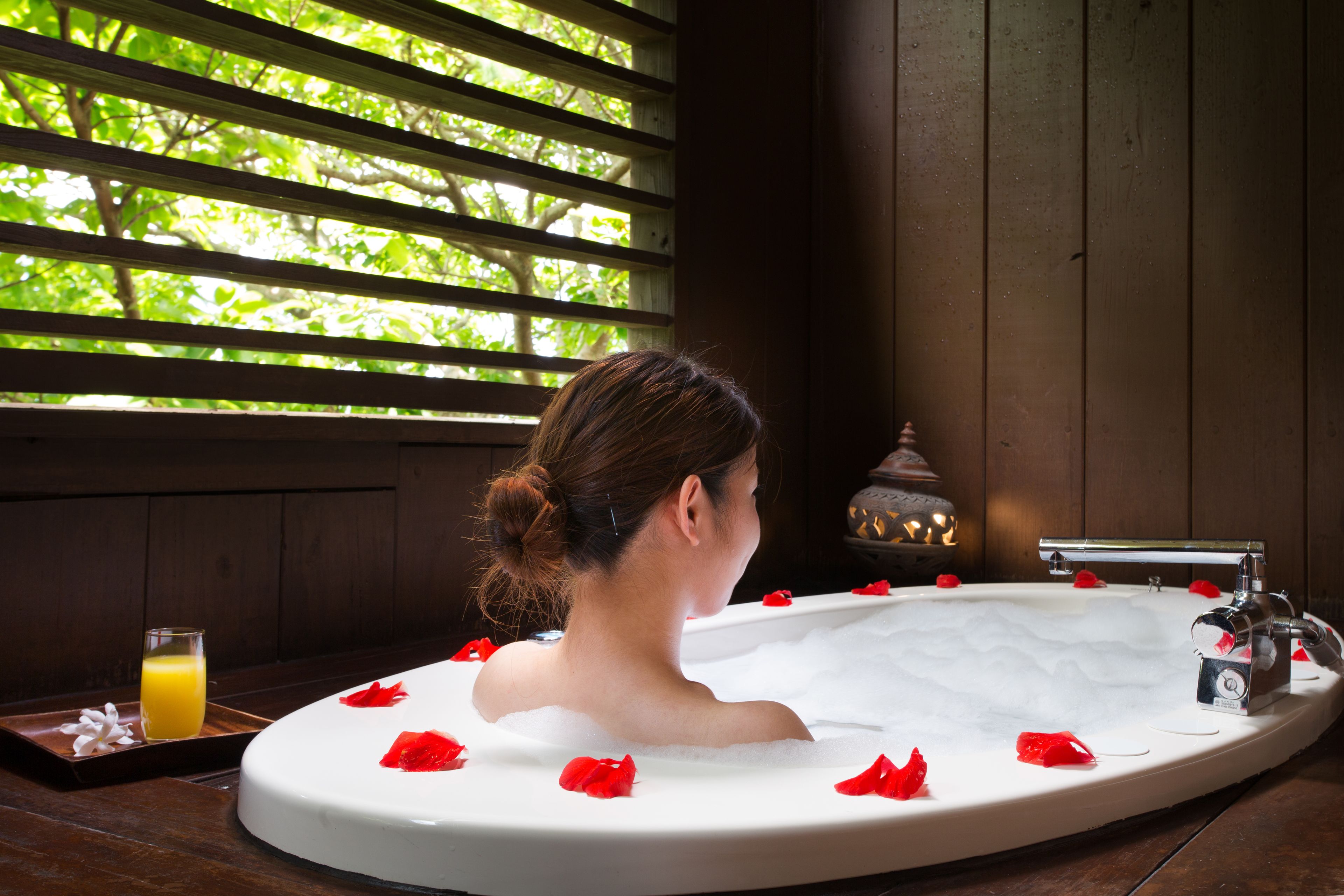 A woman in a bathtub with rose petals, a glass of orange juice, and a candle on a wooden table.