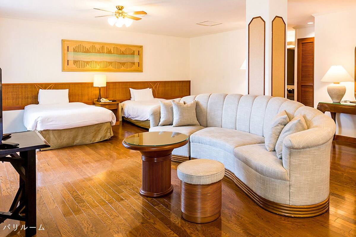 A hotel room with a large L-shaped sofa, two single beds, a round coffee table, and a small ottoman.