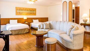 Una habitación de hotel con un amplio sofá en forma de L, dos camas individuales, una mesa de centro redonda y un pequeño otoman.