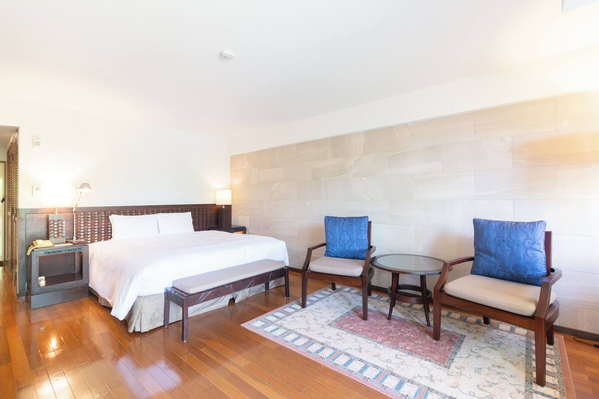 A hotel room with a bed, two armchairs, a small table, and a nightstand with a lamp.
