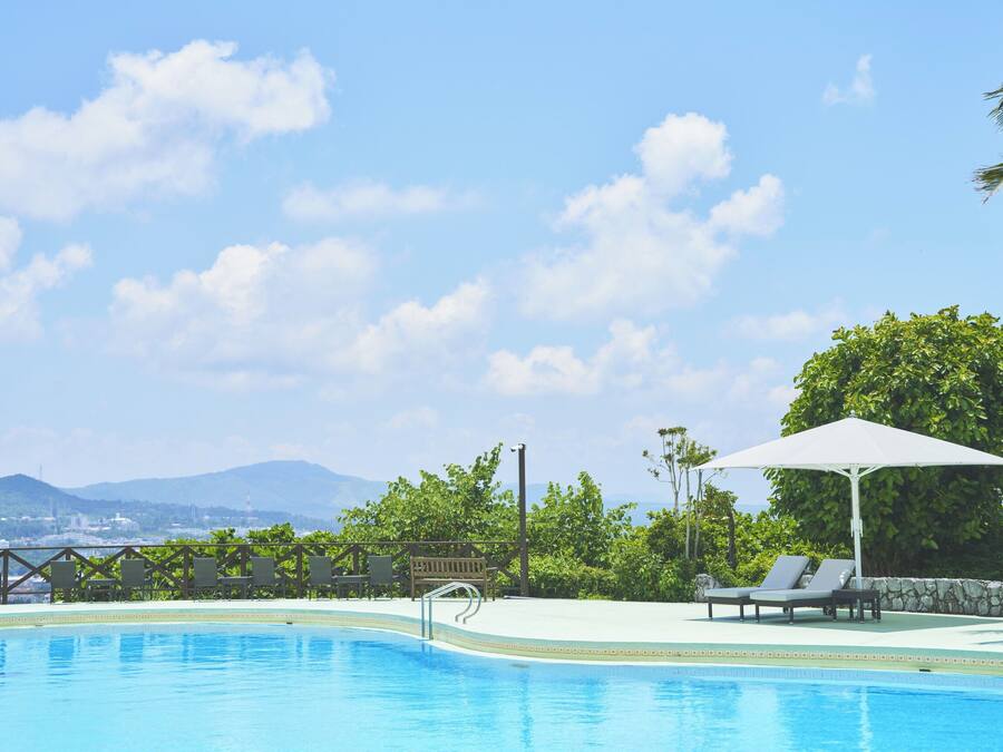 Piscina com espreguiçadeiras e guarda-sóis, cercada por vegetação e montanhas ao fundo.