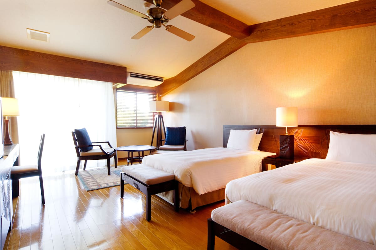 A hotel room with two beds, a desk, a chair, and a ceiling fan.