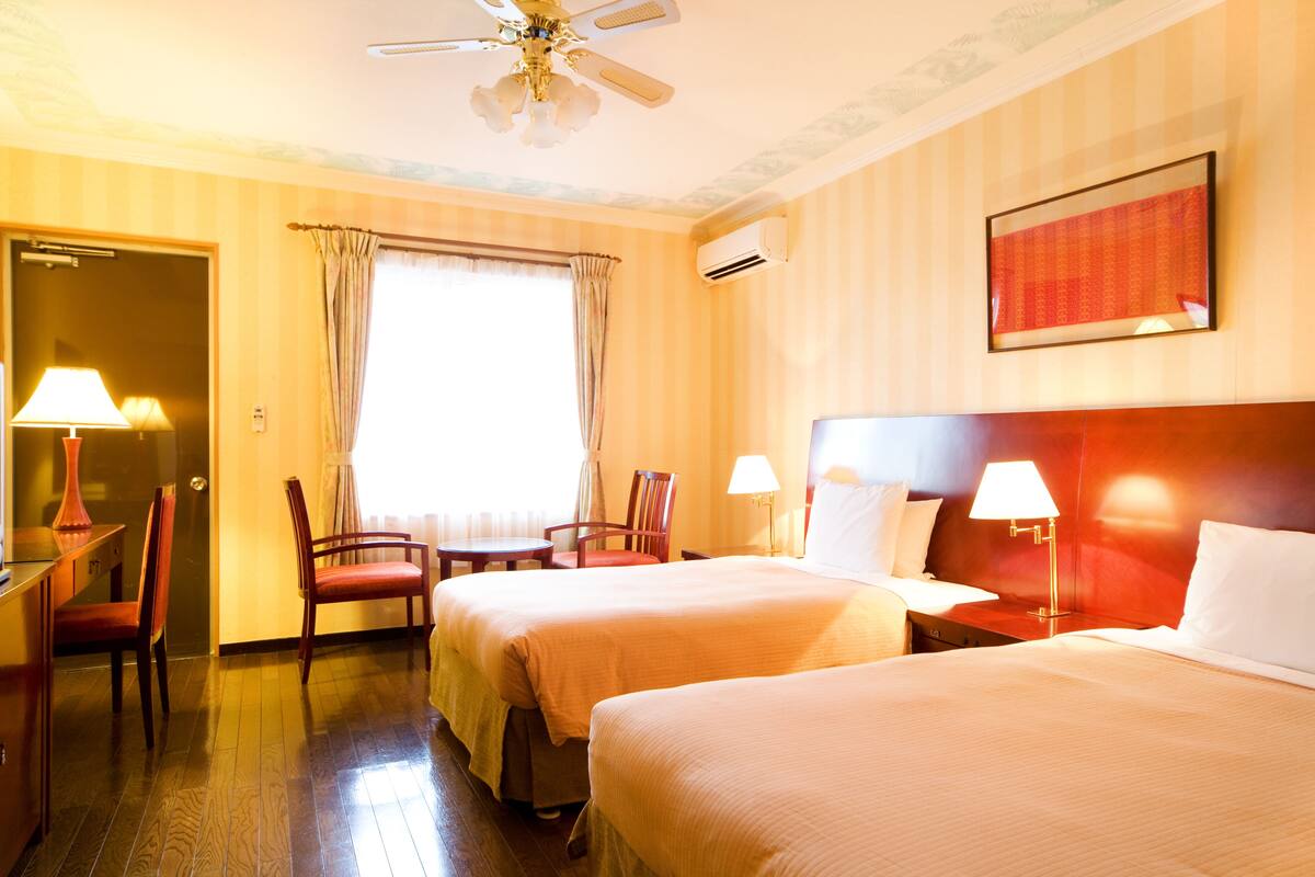A hotel room with two beds, a desk with a lamp, a chair, a window with curtains, and a ceiling fan.