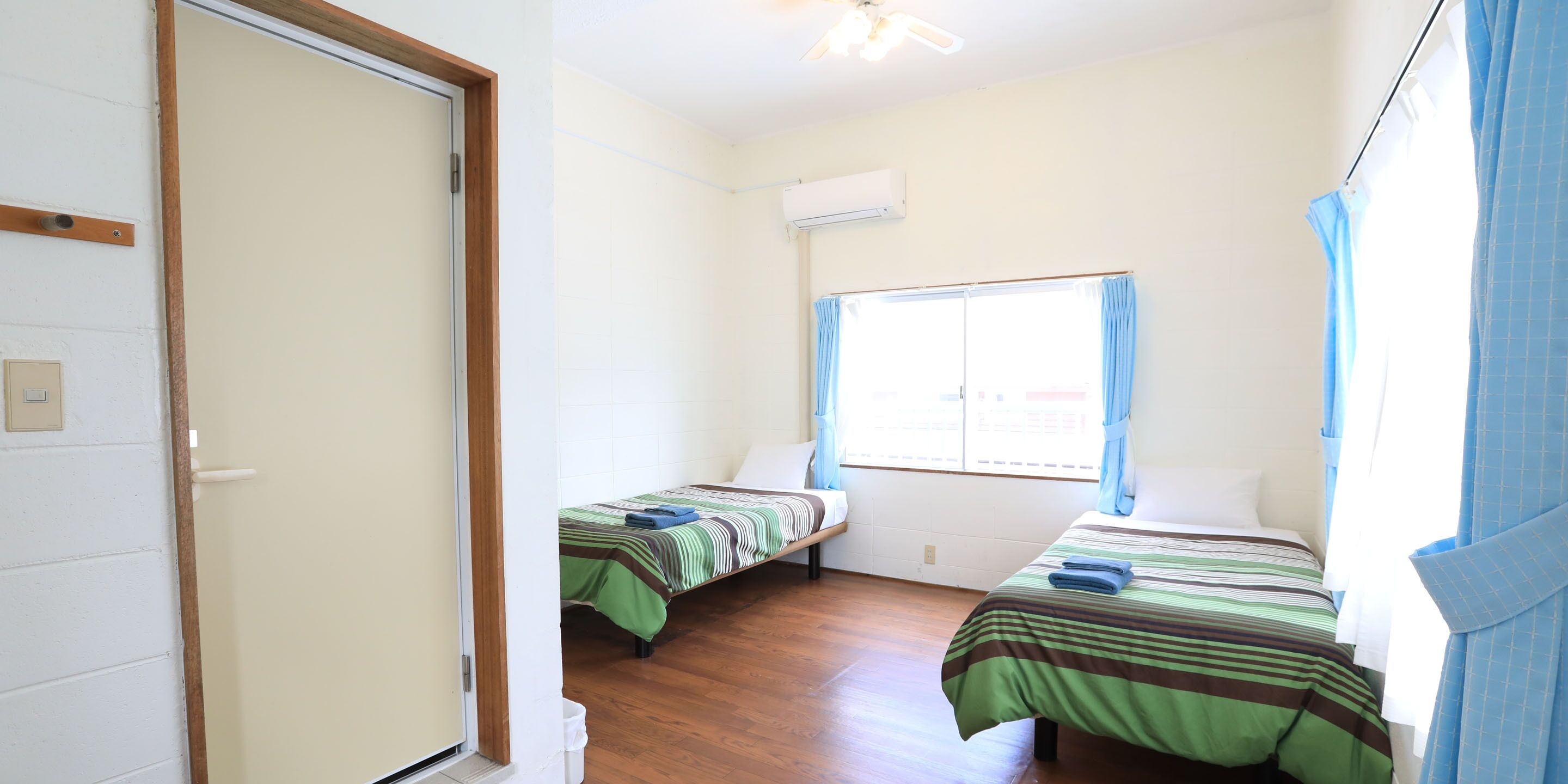A room with two single beds, a window with curtains, an air conditioning unit, and a wooden floor.