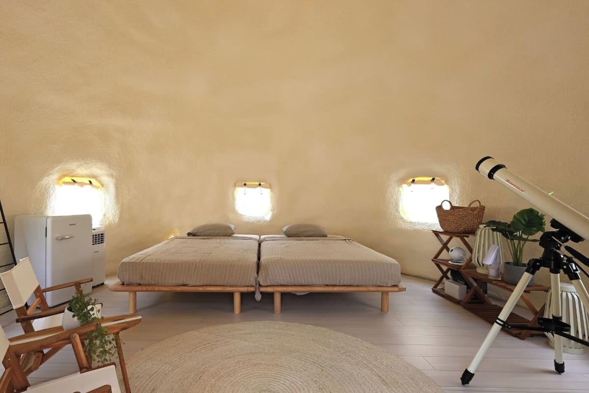 Un dormitorio con cama, mesitas de noche, lámpara, un trípode y una ventana.