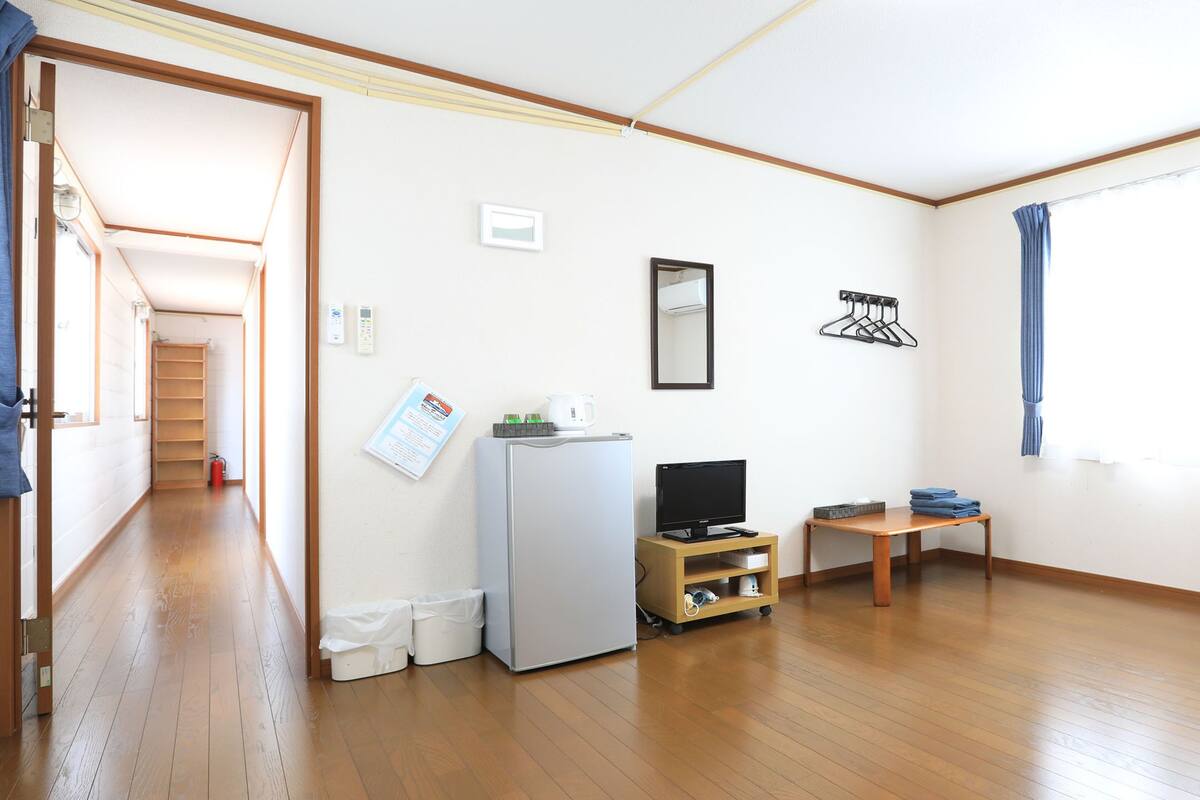 Habitación con piso de madera, cama con almohadas, una mesita, televisor, refrigerador y ventana con cortinas.
