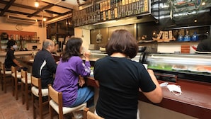 Un interior de restaurante con sillas y mesas de madera, una barra con vitrina y un cartel con el menú encima de la barra.