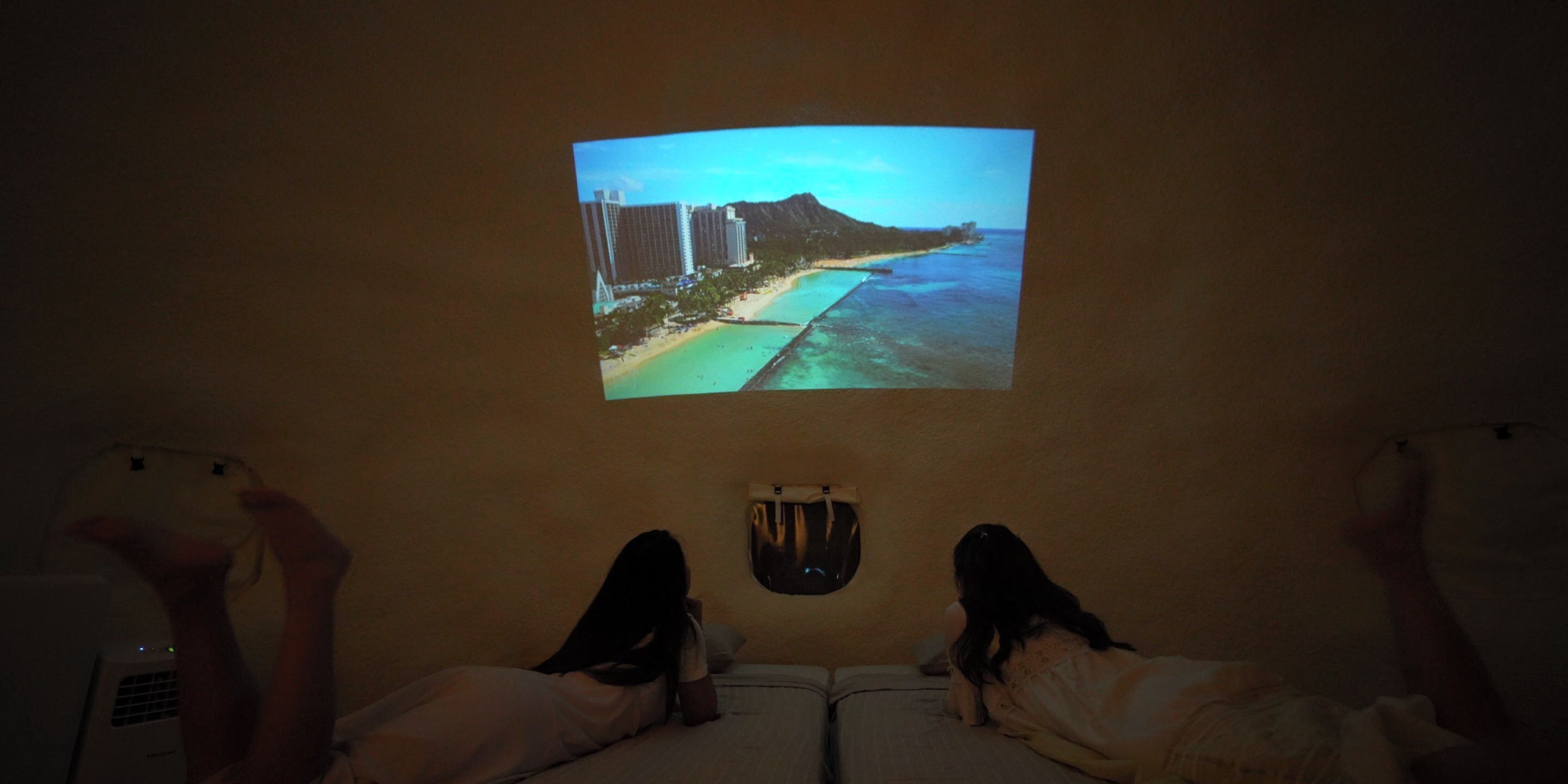 Une projection représentant une scène de plage sur un mur, deux lits avec des oreillers et une petite fenêtre.