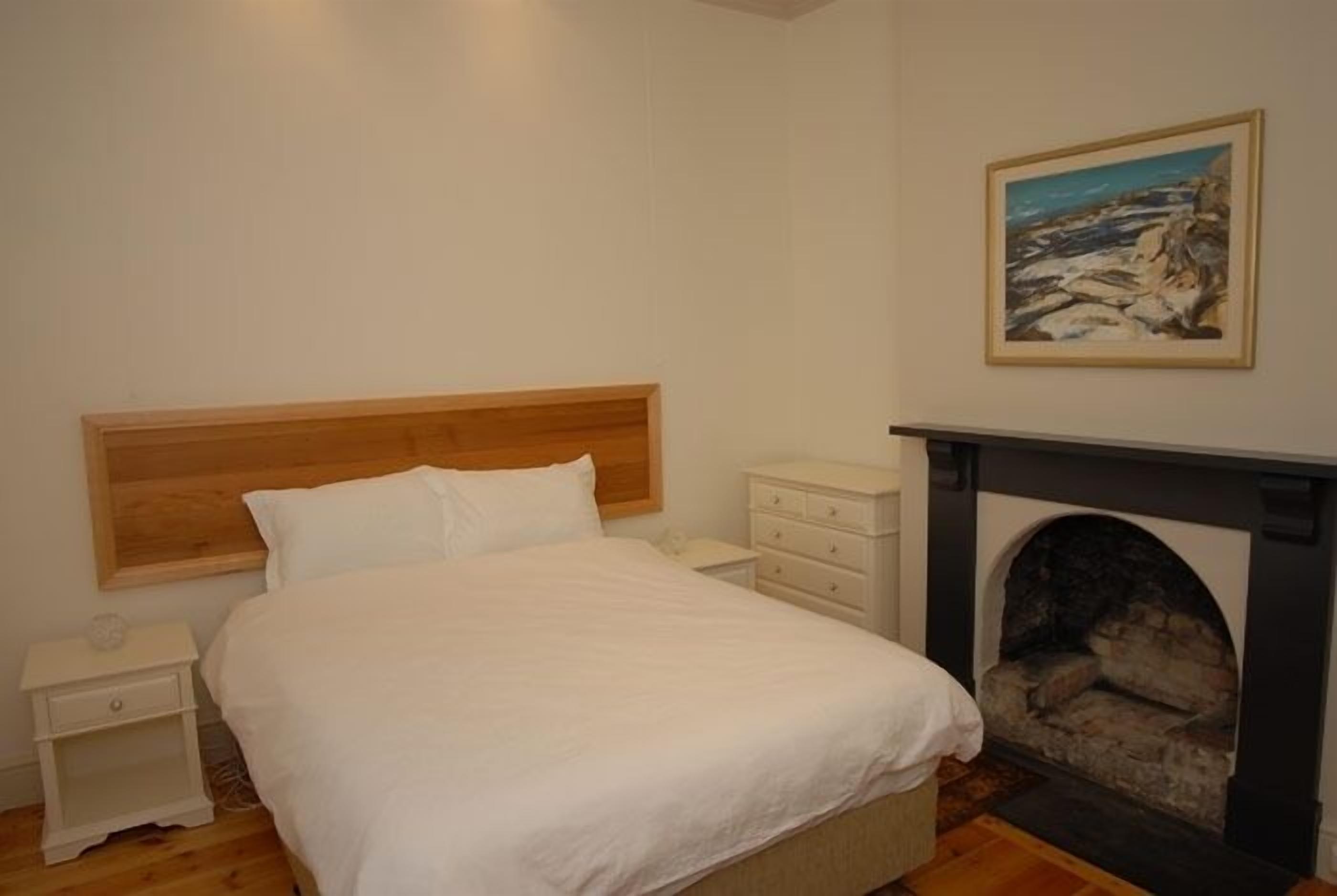 Ein Schlafzimmer mit hölzernem Kopfteil, einem weißen Bett, einem Nachttisch, einer Kommode und einem Kamin.