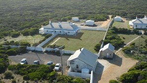 Vue aérienne d’une propriété comprenant plusieurs bâtiments, dont une grande maison blanche à toit en pente et plusieurs constructions plus petites. On y voit une allée pavée et un espace de stationnement avec des voitures.