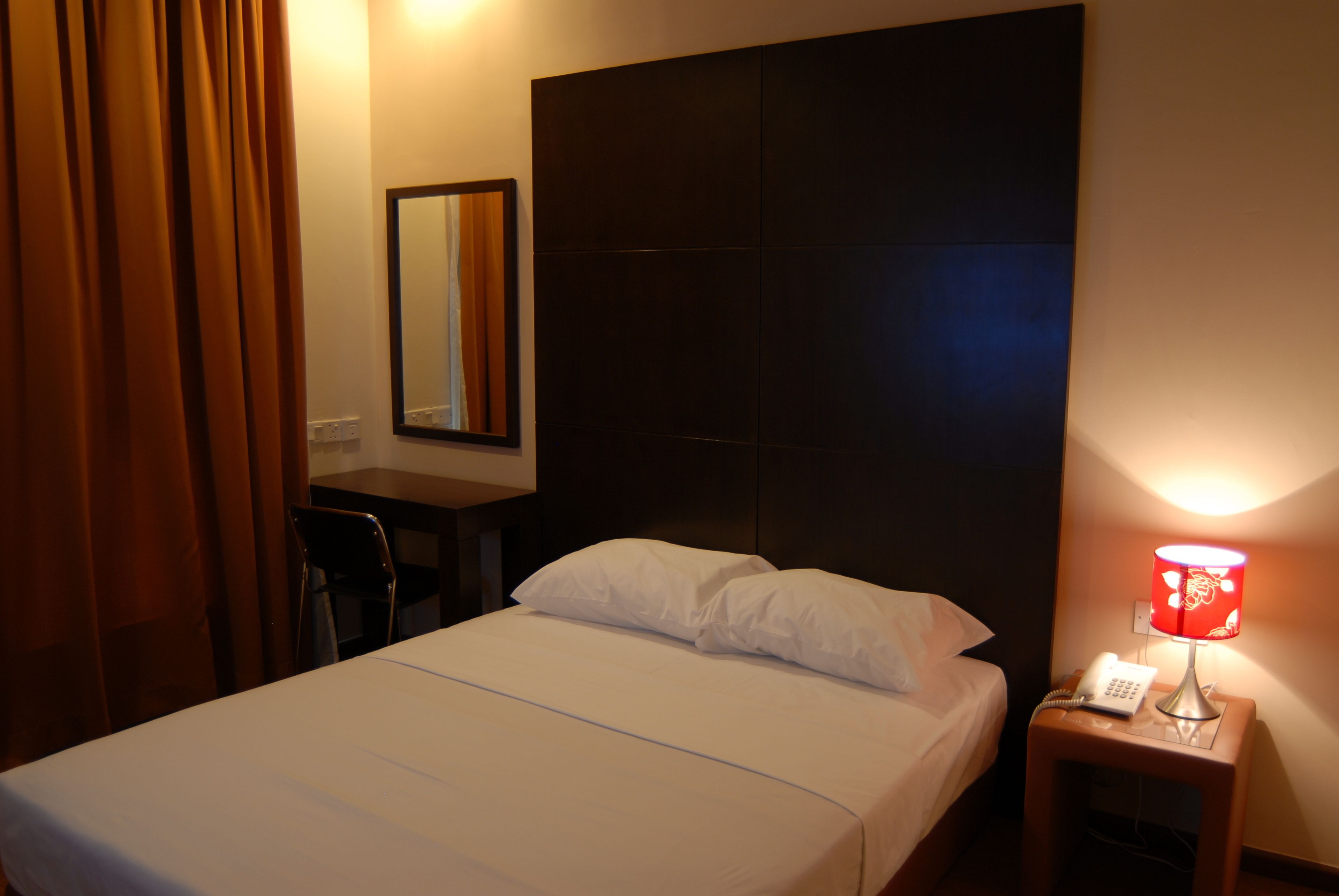 Habitación de hotel con una cama, un escritorio, una silla, una mesita de noche con lámpara y un espejo en la pared.