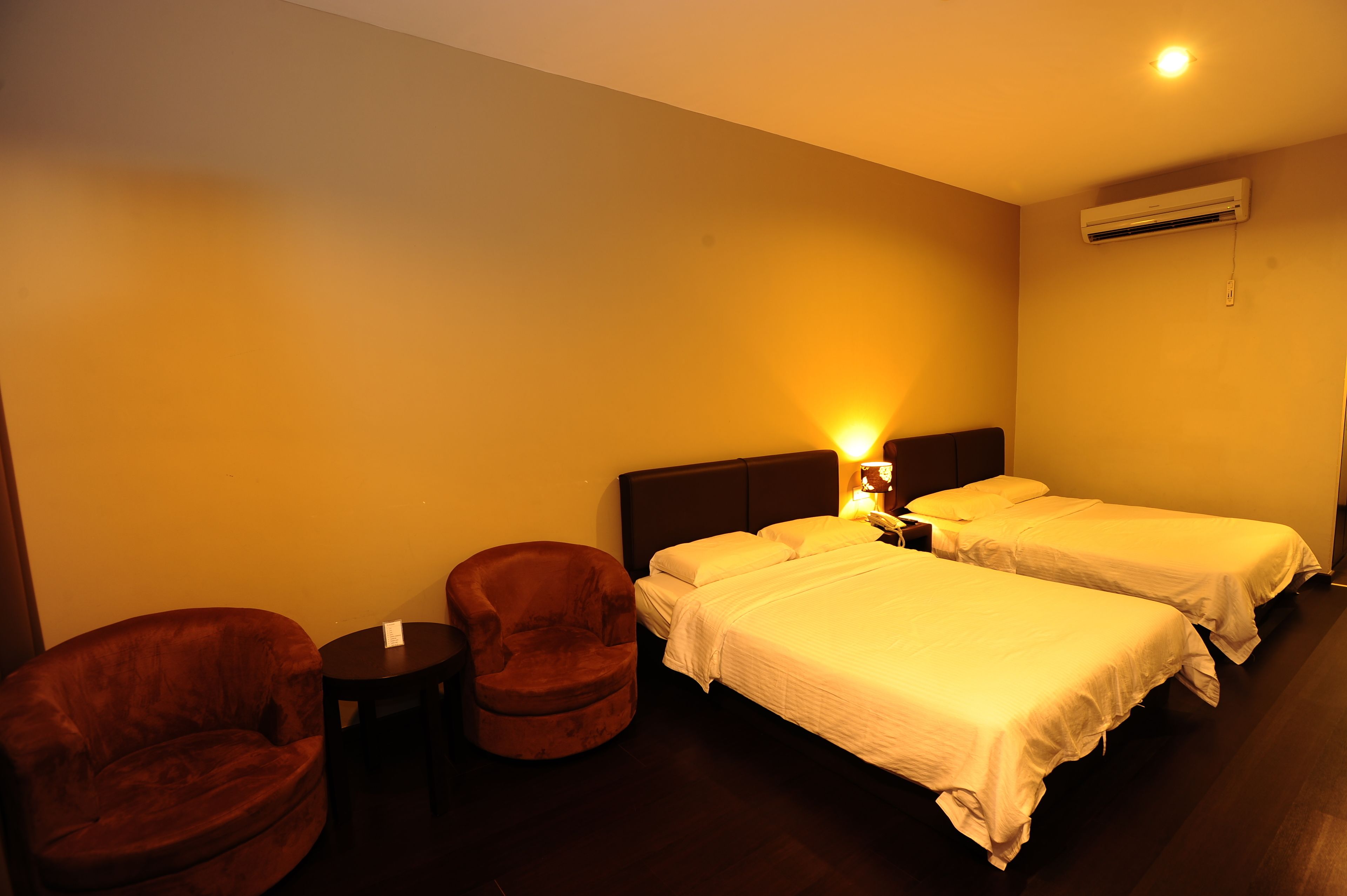 Une chambre d’hôtel avec deux lits simples, un fauteuil marron, une petite table ronde et une table de chevet avec une lampe.