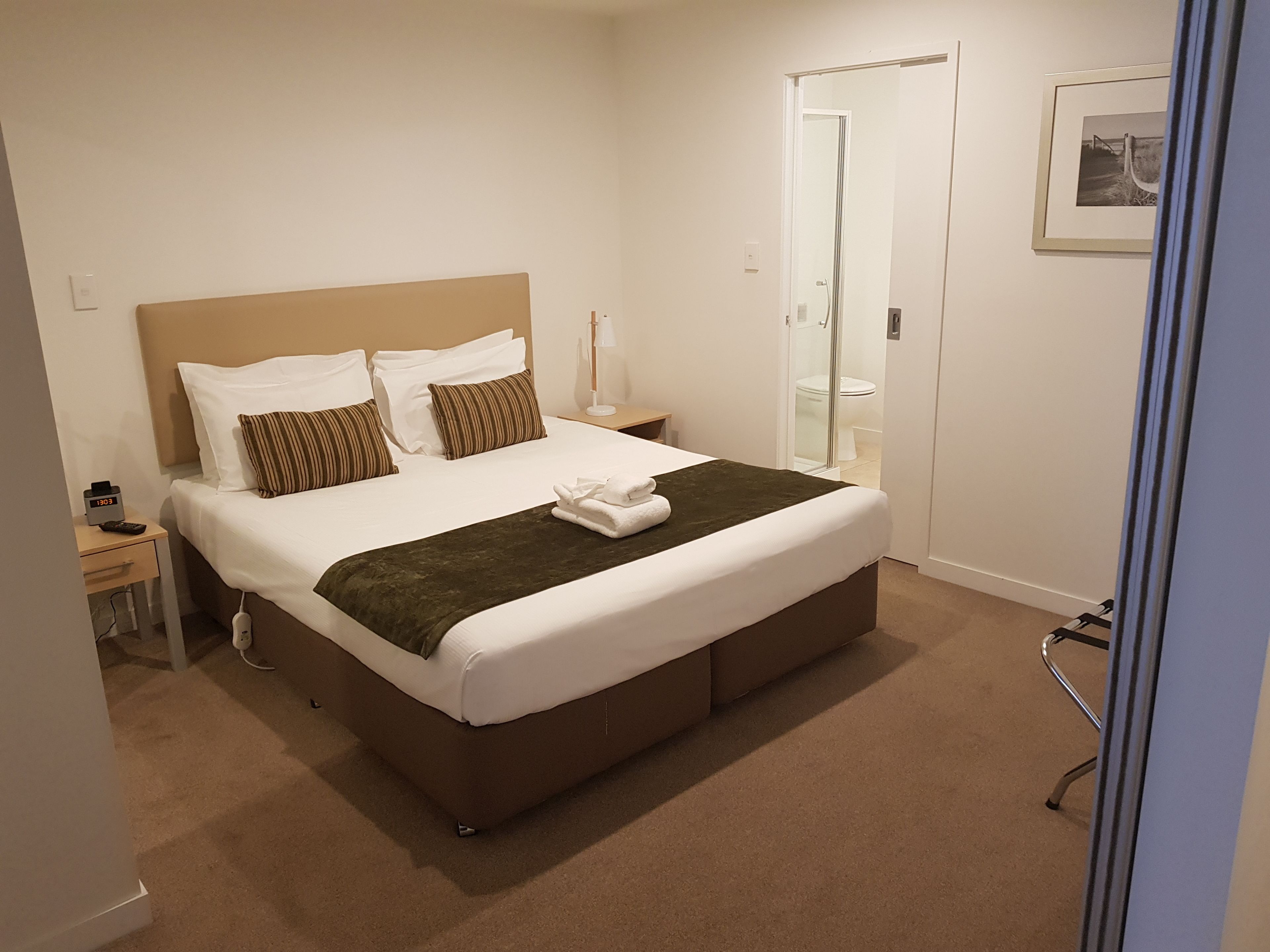 Een netjes opgemaakt bed met witte lakens en een bruin hoofdeinde, een nachtkastje met een lamp, en een ingelijst beeld aan de muur.