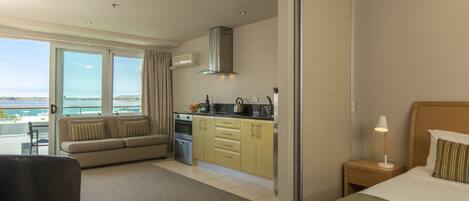 A modern hotel room with a large bed, a sofa, a kitchenette, and a view of the sea.
